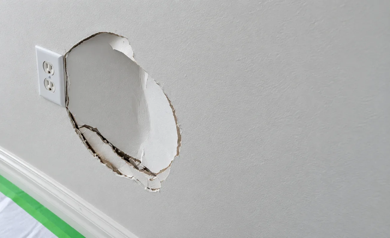 Large broken hole in a drywall wall beside an electrical outlet, taped off and ready for patching by AllPro AG in San Diego