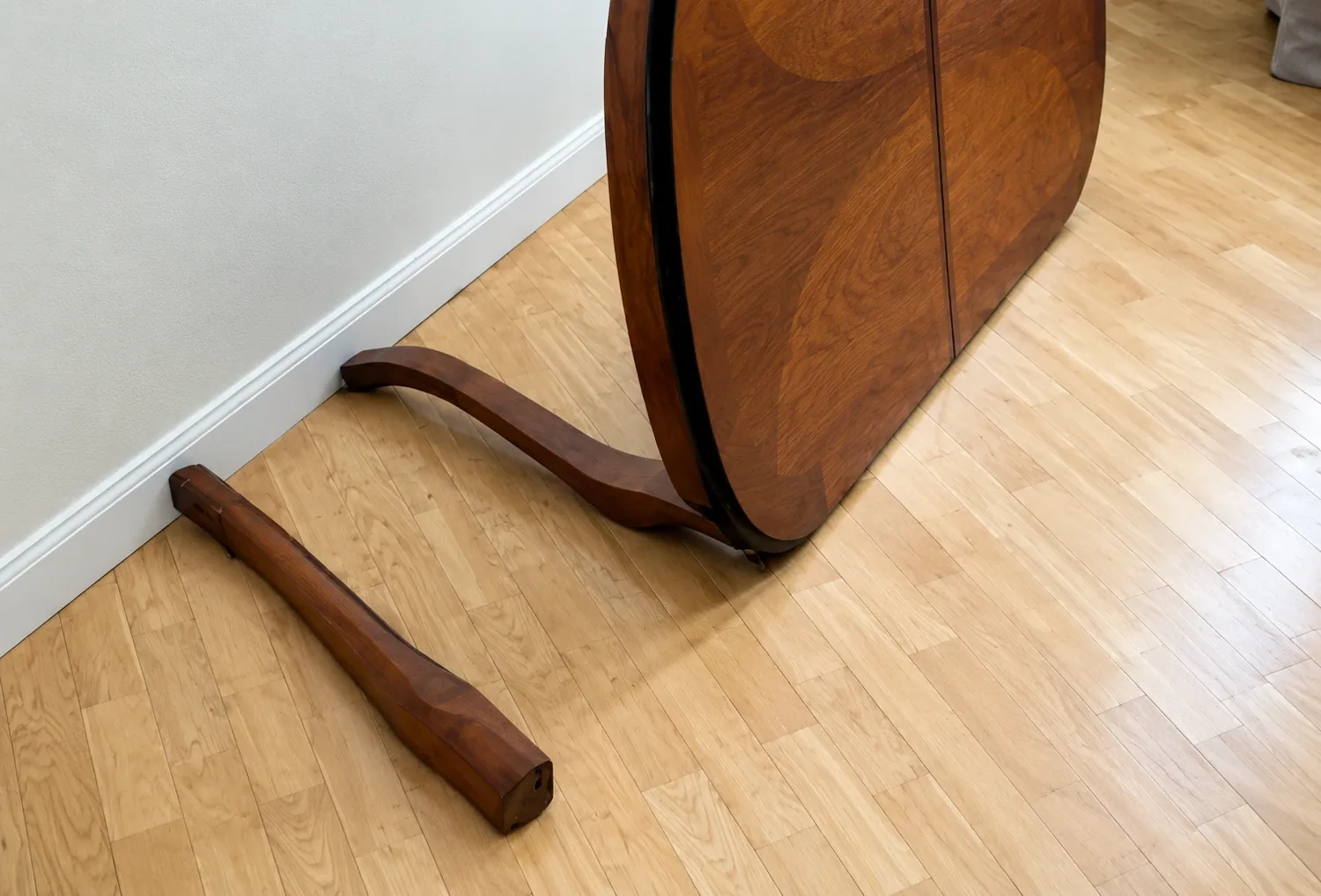 Round walnut dining table tipped on its side with a curved leg snapped off and lying on the hardwood floor, a common furniture repair in San Diego